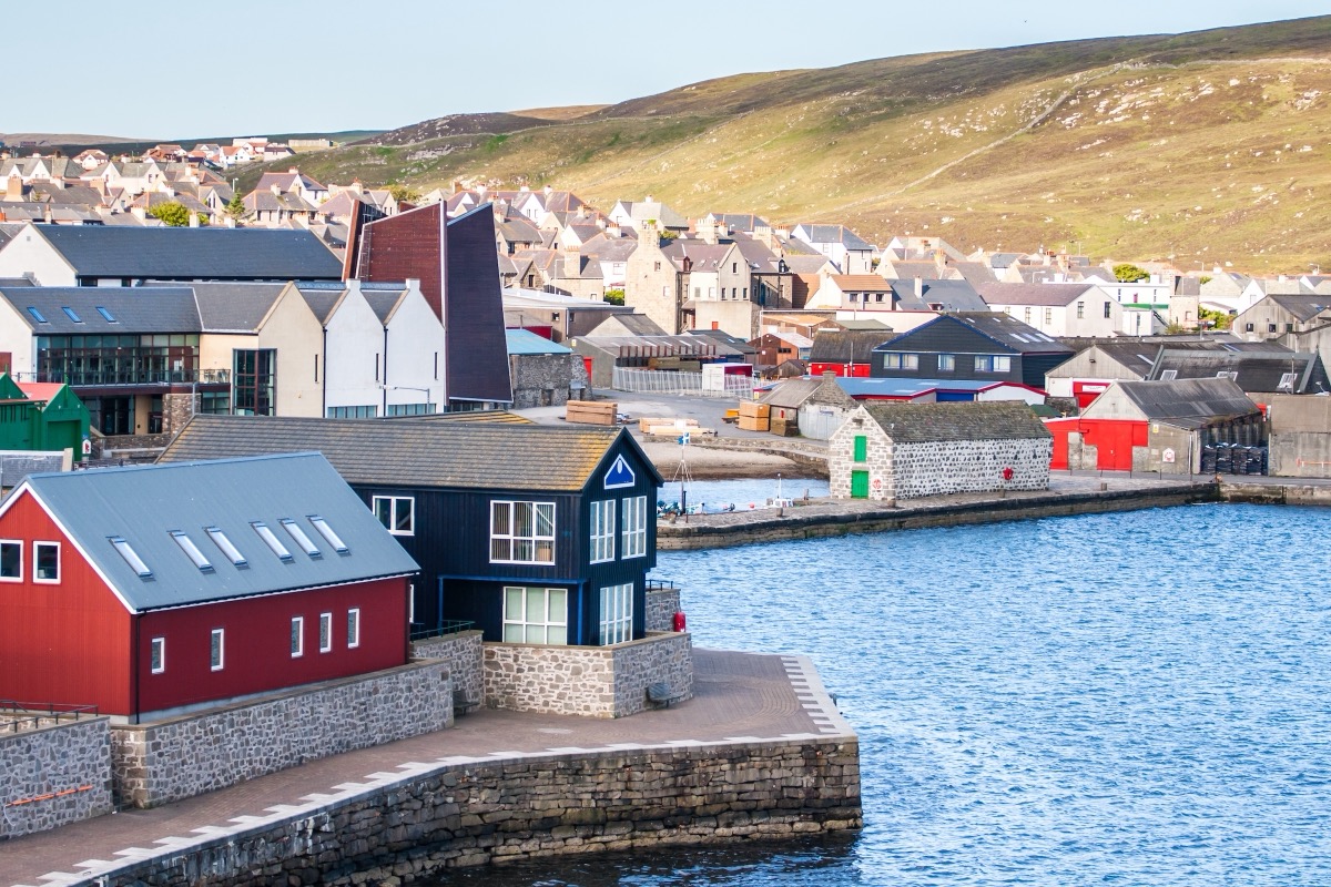 Colorful warehouses and houses along the quay of Lerwick in the Shetland Islands, with the harbor and gentle green hills behind