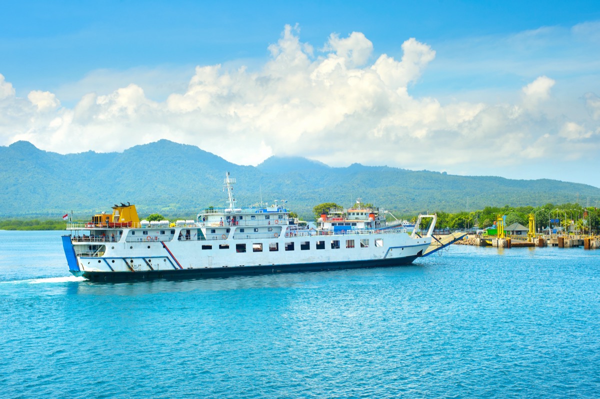 A white ferry on the Bali–Java route leaving the port, with tropically forested hills and port buildings in the background