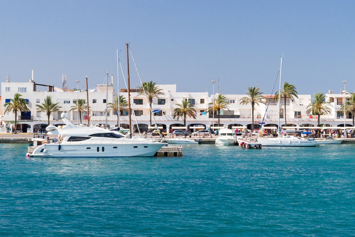 Der Hafen von La Savina auf Formentera mit Yachten und Ausflugsbooten am Kai, Palmenreihe und weißen Hafengebäuden über türkisfarbenem