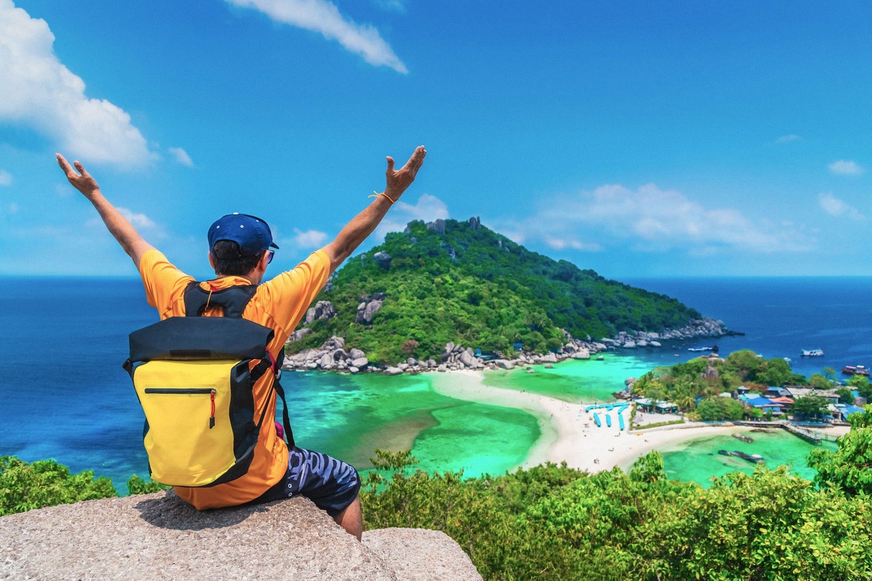 A traveller with a backpack sits on a rocky outcrop, arms raised, looking out