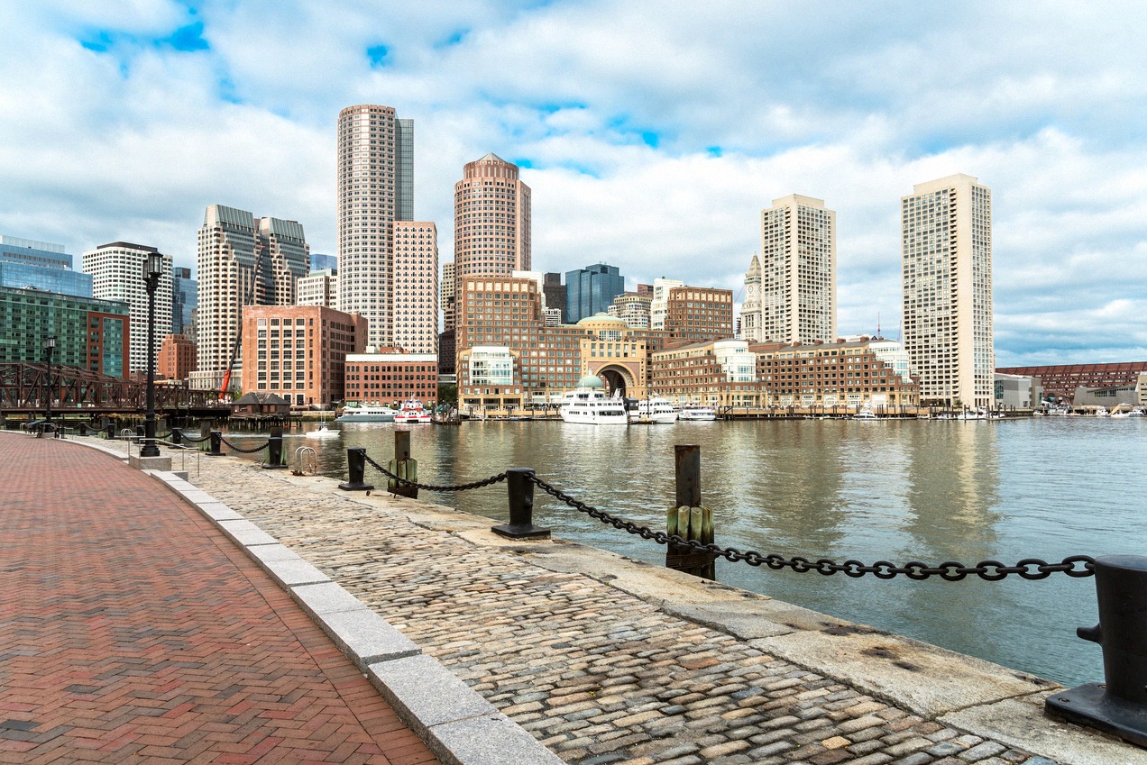 The Boston skyline seen from a paved harbour walk with ferry landings and calm water
